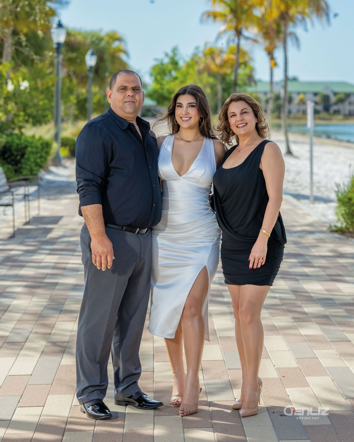 Family of three posing outdoors on a paved walkway with palm trees and waterfront in the background.