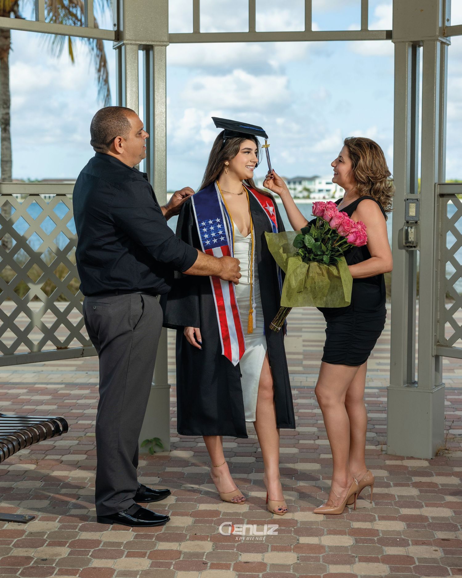 Graduate in cap and gown poses with proud parents holding flowers at an outdoor pavilion.