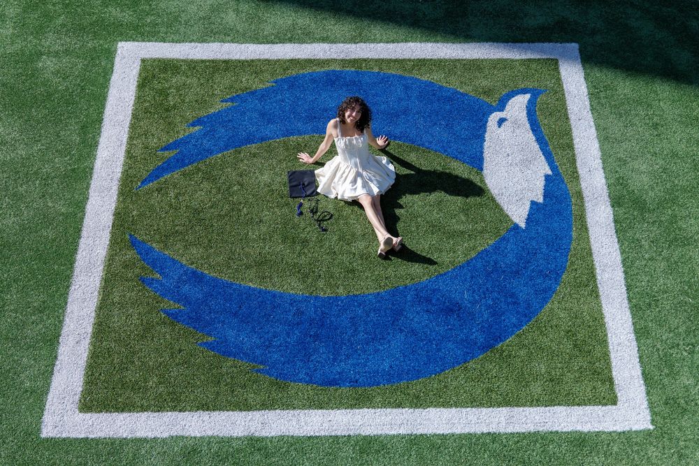 Aerial view of graduate in white dress lying on blue school logo on turf field.