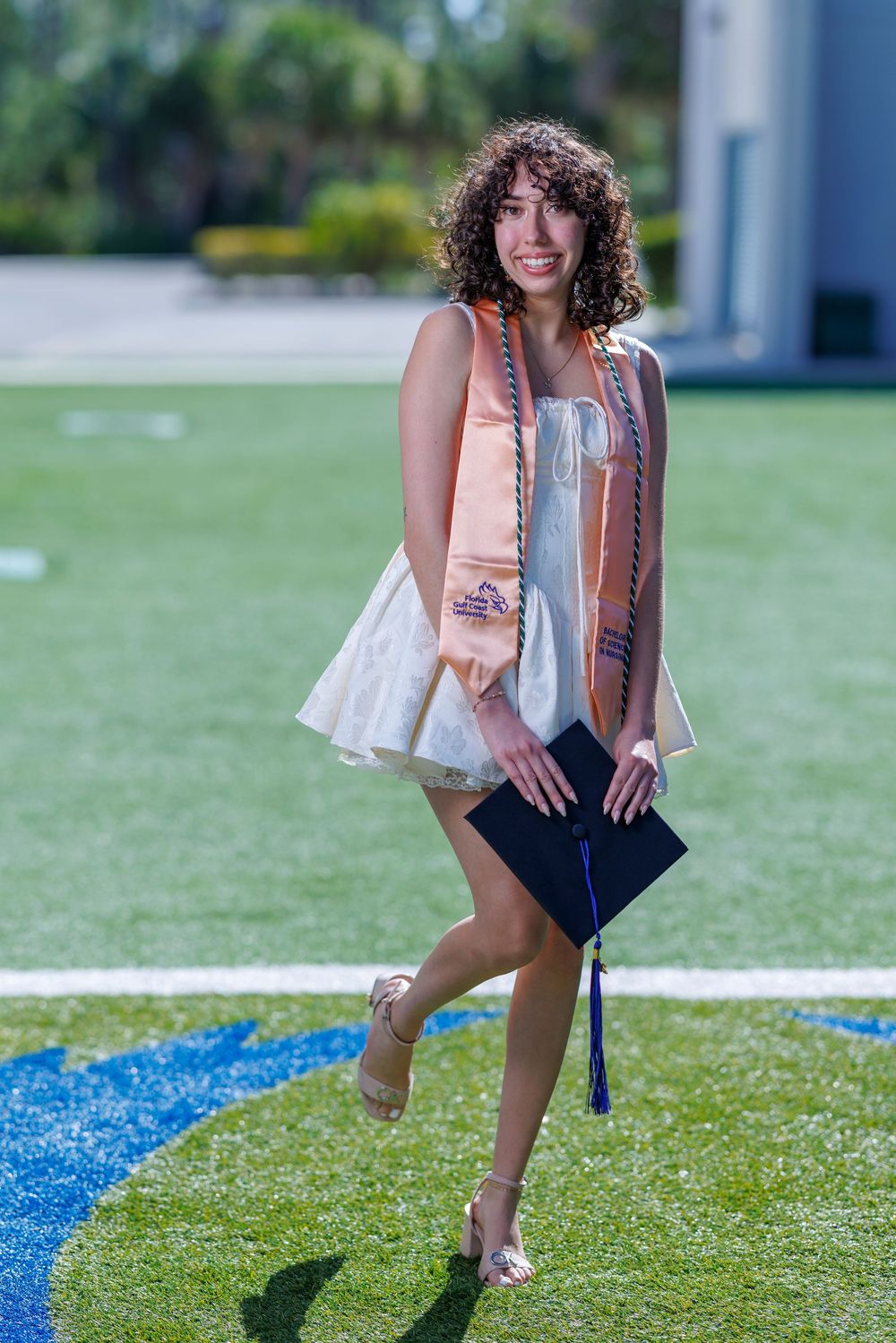 Smiling young woman in white dress holding graduation cap on green field with honor stoles.