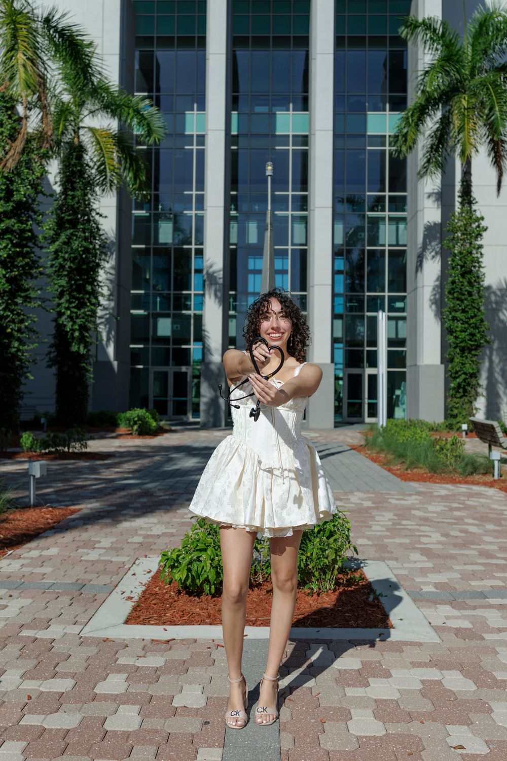 Young woman in white dress holding stethoscope in front of modern building with palm trees.