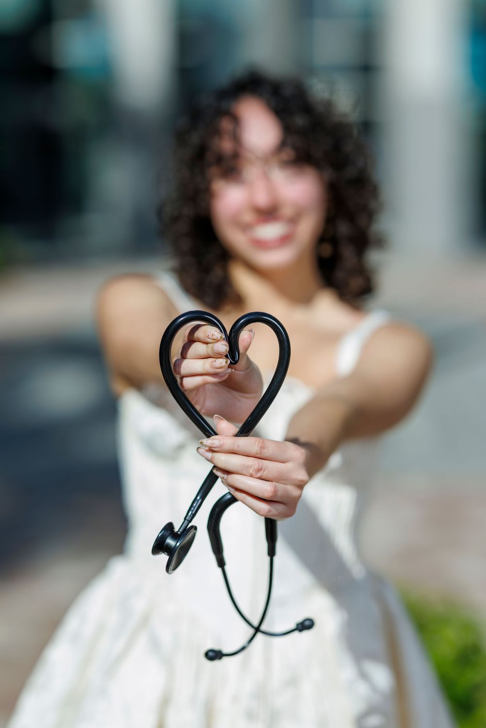 Smiling graduate holding black stethoscope shaped into heart toward camera outdoors.