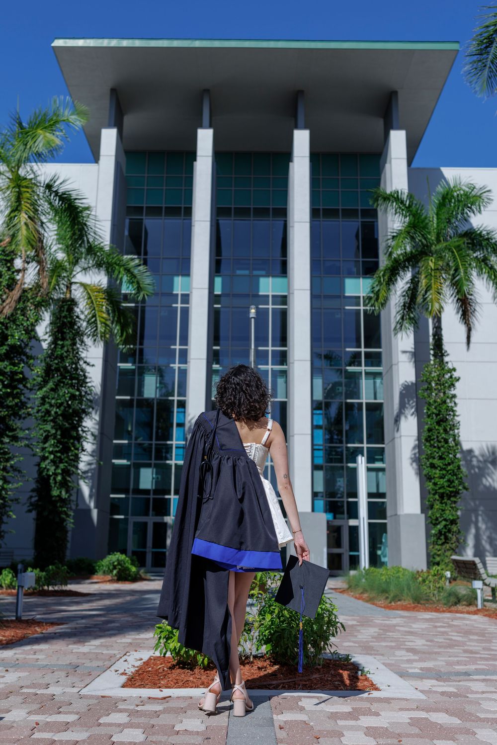 Graduate in white dress and black gown walking toward modern university building with palms.