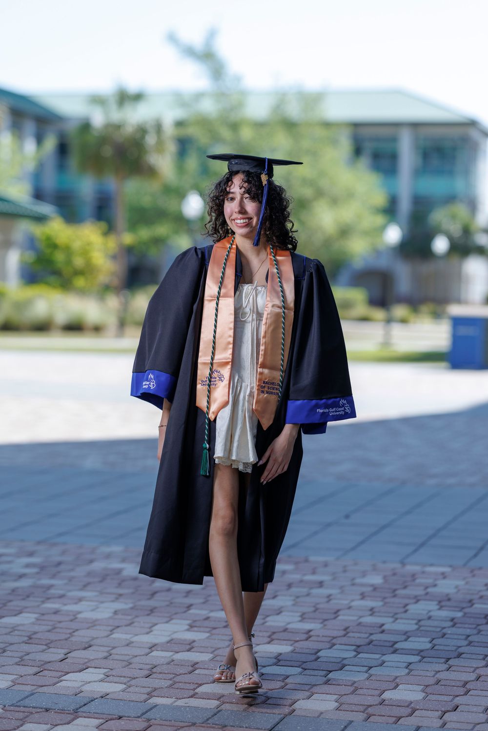 Young woman in black graduation gown and cap with orange stole walks on brick path at FGCU campus.