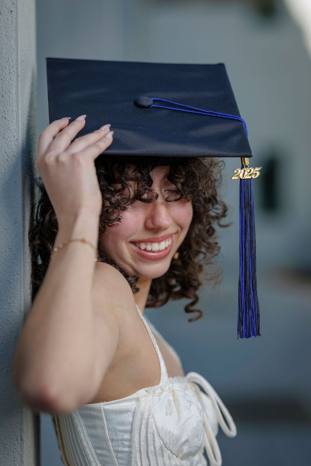 Joyful curly-haired graduate smiles while holding her 2025 navy blue mortarboard cap outdoors.