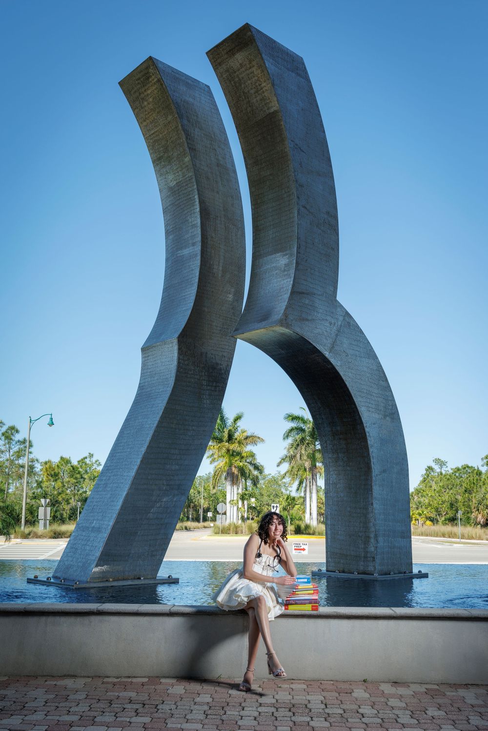 Graduate in white dress sits reading colorful books beside large abstract sculpture with palm trees.