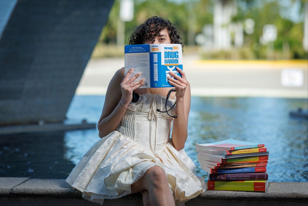Smiling graduate in white dress holds drug handbook book while sitting beside stacked textbooks outdoors.