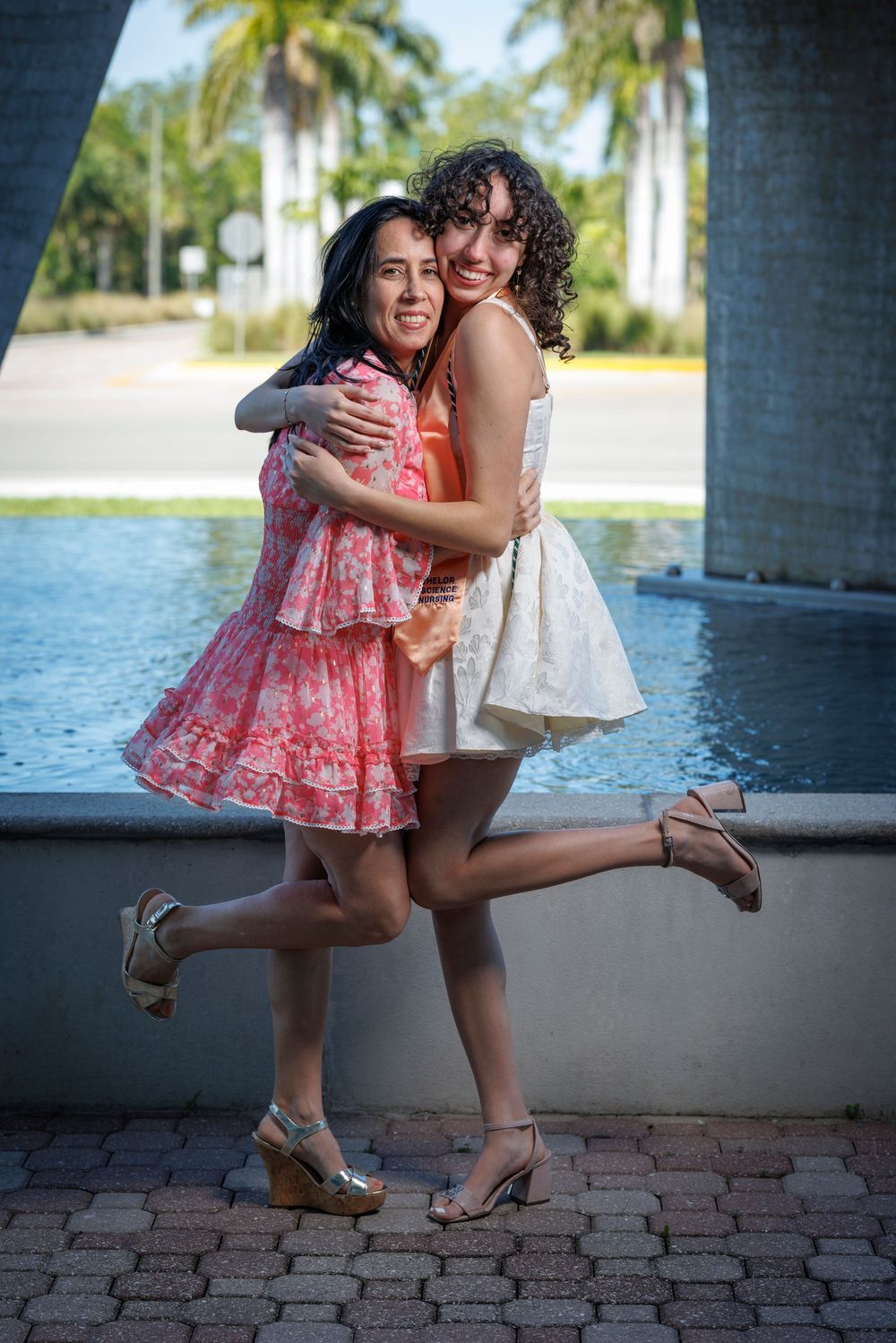 Two young women in colorful dresses embrace and laugh joyfully near a fountain with palm trees.