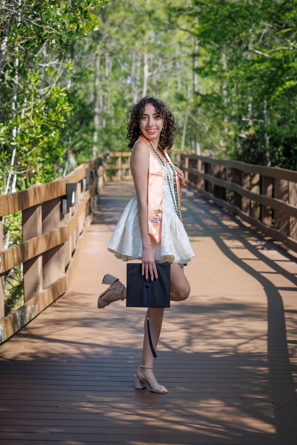 Smiling graduate in white dress holds cap while posing on wooden bridge surrounded by lush green trees.