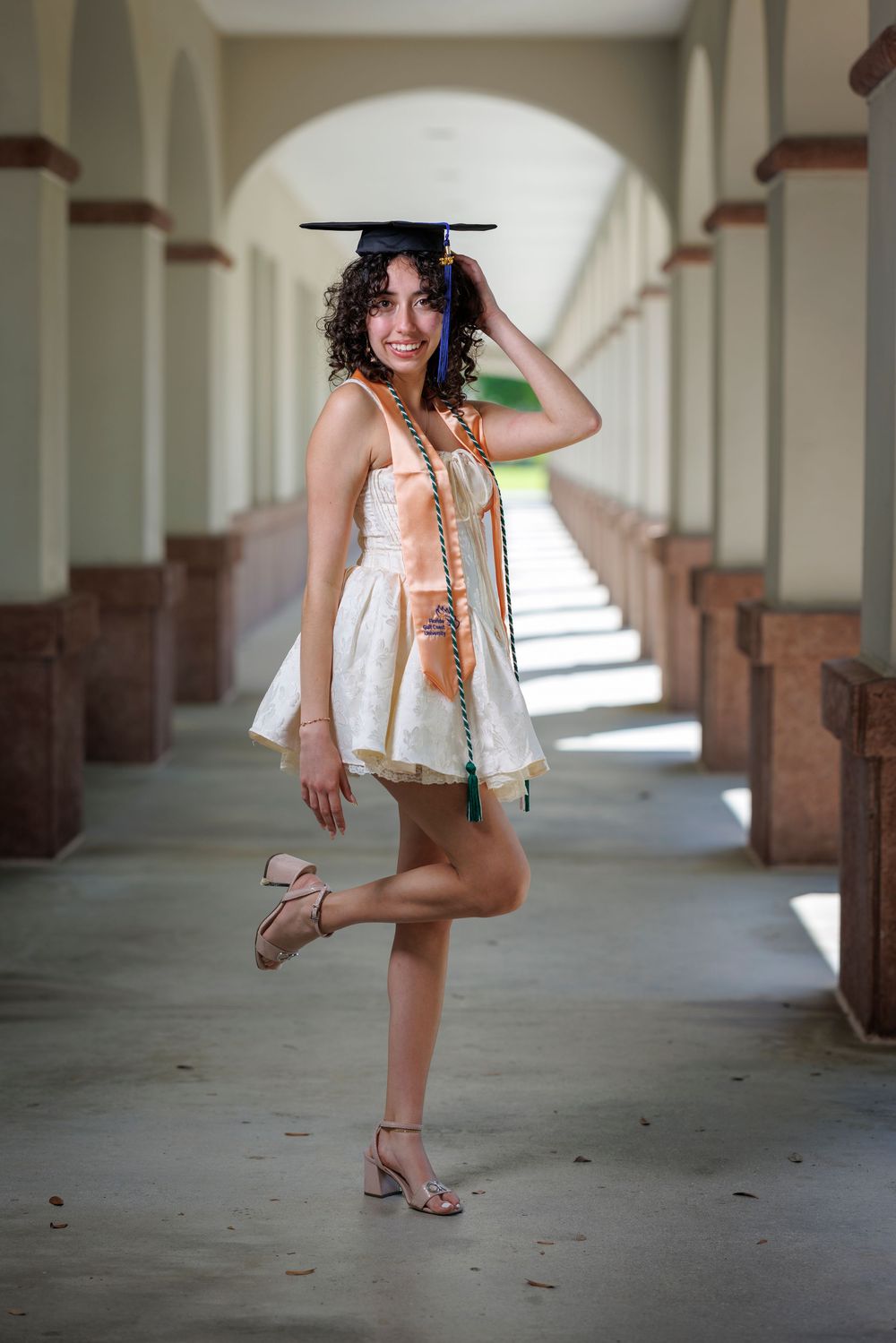 Smiling graduate in white dress and cap with honor cords poses playfully in arched campus corridor.