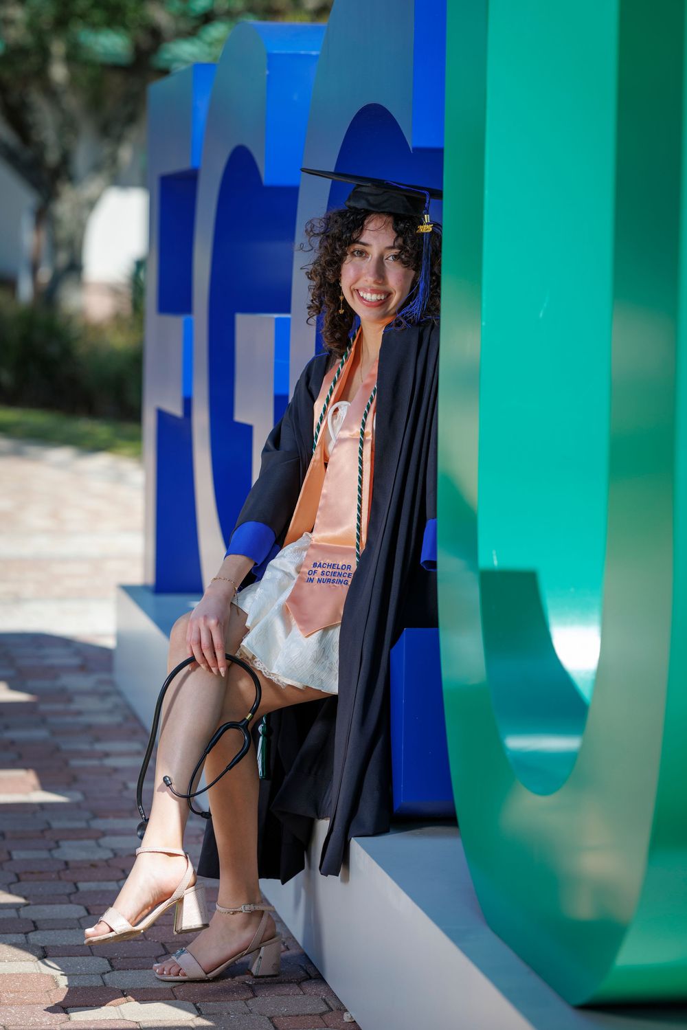 Smiling graduate in black gown and cap sits relaxed against large FGCU letter sign on campus.