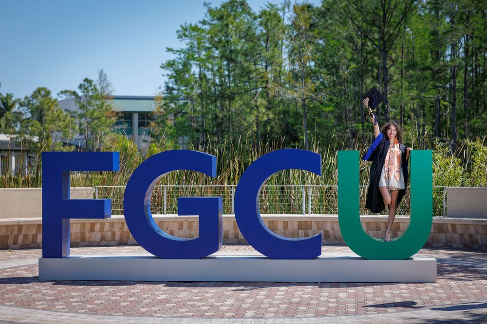 Graduate in cap and gown celebrates beside large colorful FGCU letters sign on sunny campus grounds.