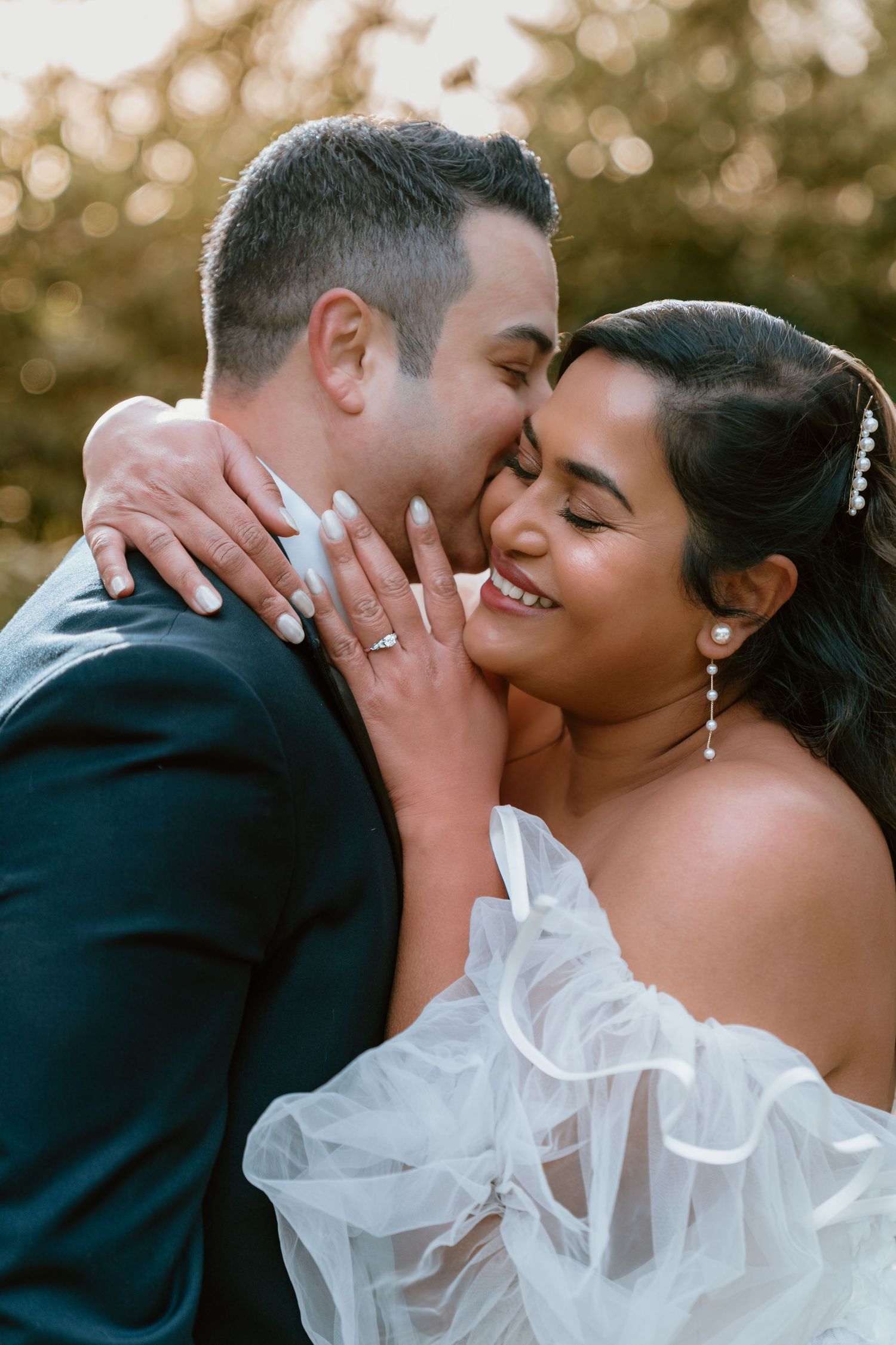 A groom kisses his bride on the cheek as she smiles, both dressed in wedding attire outdoors.