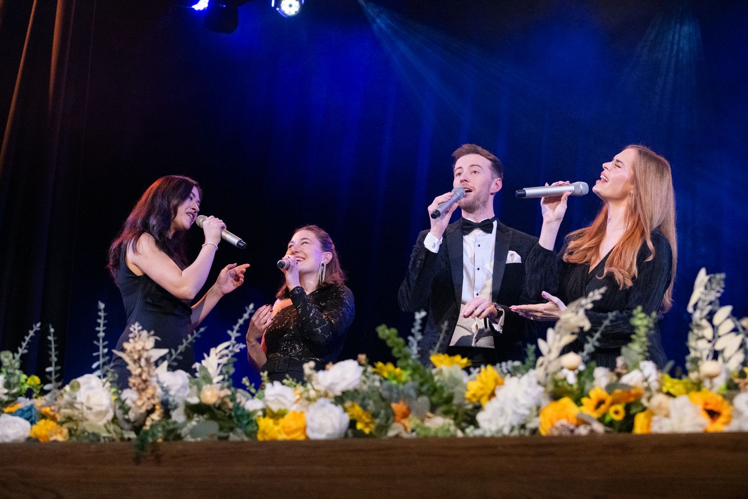 Four performers singing on a floral-decorated stage under blue lights at a formal event.