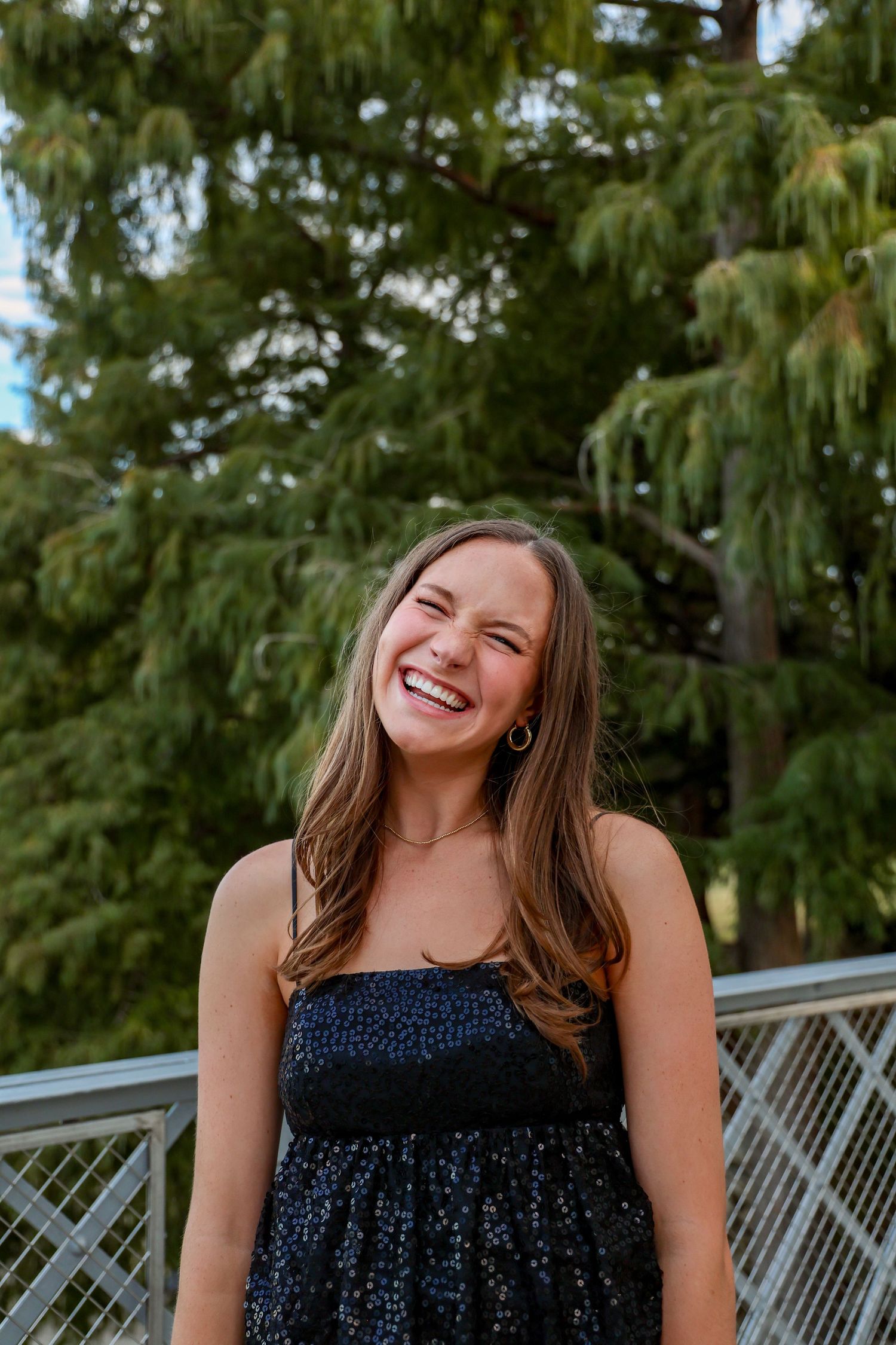 A young woman laughing joyfully outdoors in a black sequin dress with green trees in the background.