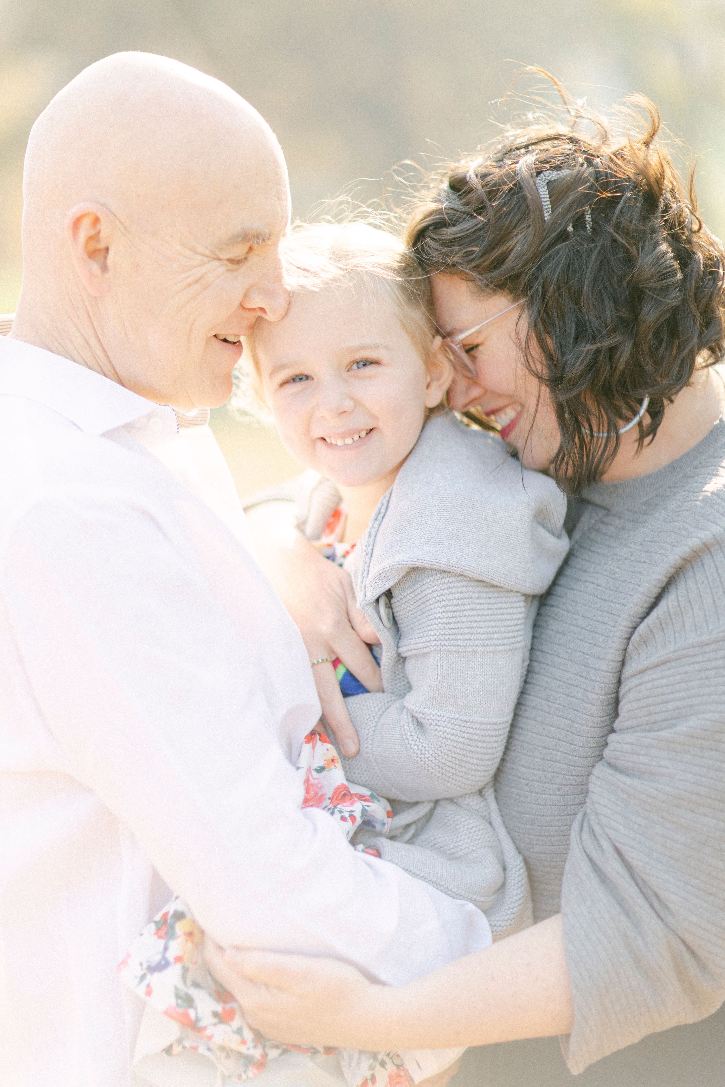 A loving family embraces in soft sunlight wearing white and grey casual clothing.