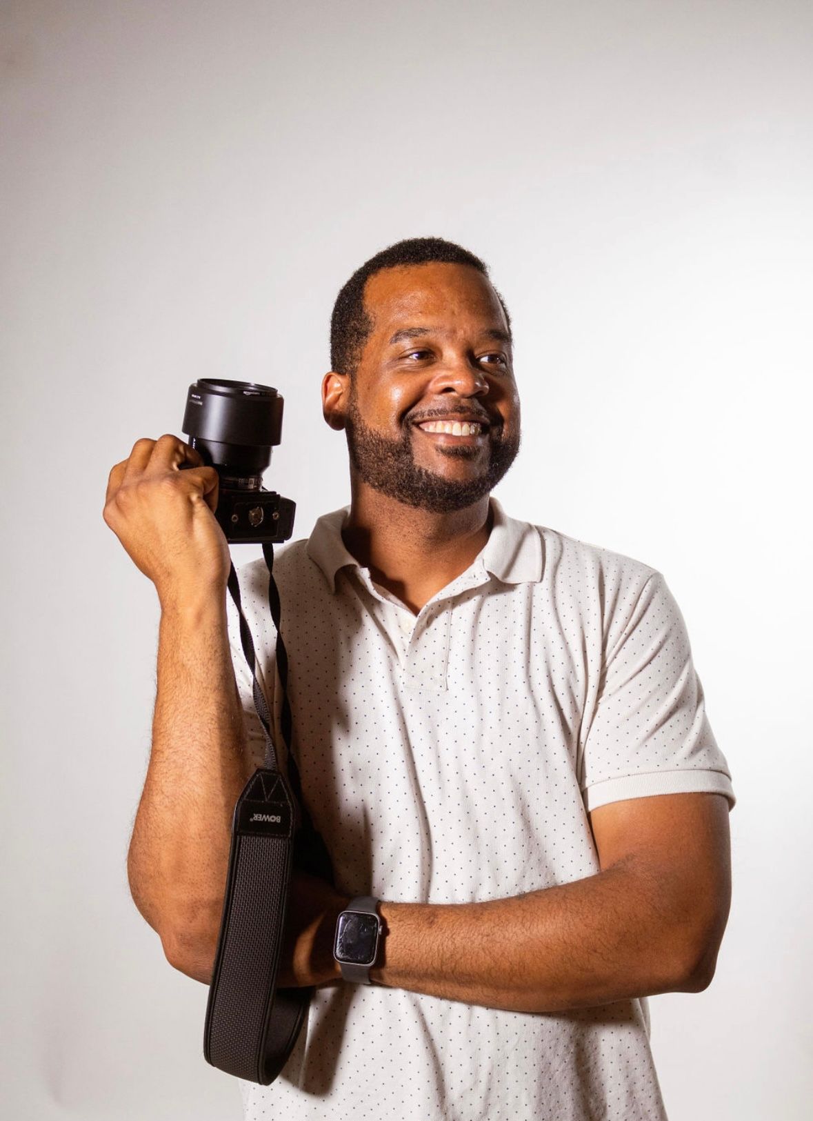 Professional photographer poses with camera equipment against white background in casual attire.