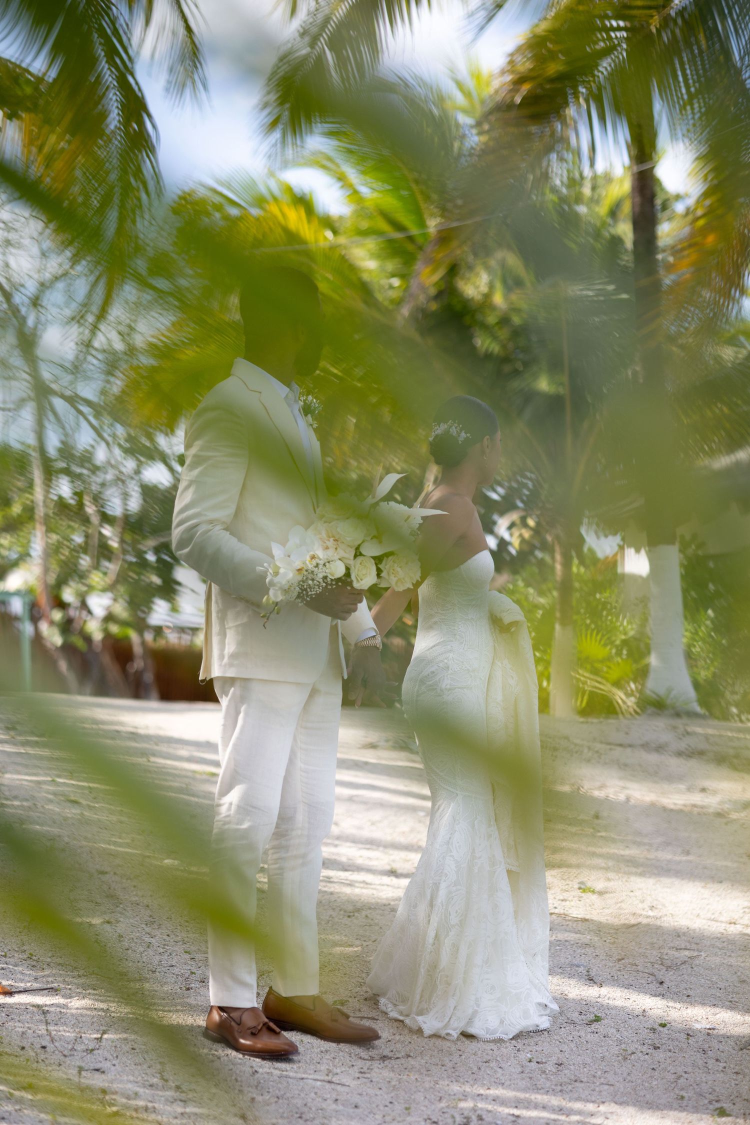 Bride and groom kissing among tropical palm trees, both dressed in white, holding a bouquet on a sandy path.