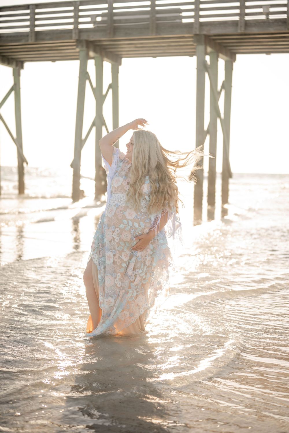 Ethereal maternity photoshoot at beach pier with flowing floral dress catching sunset light in shallow water.