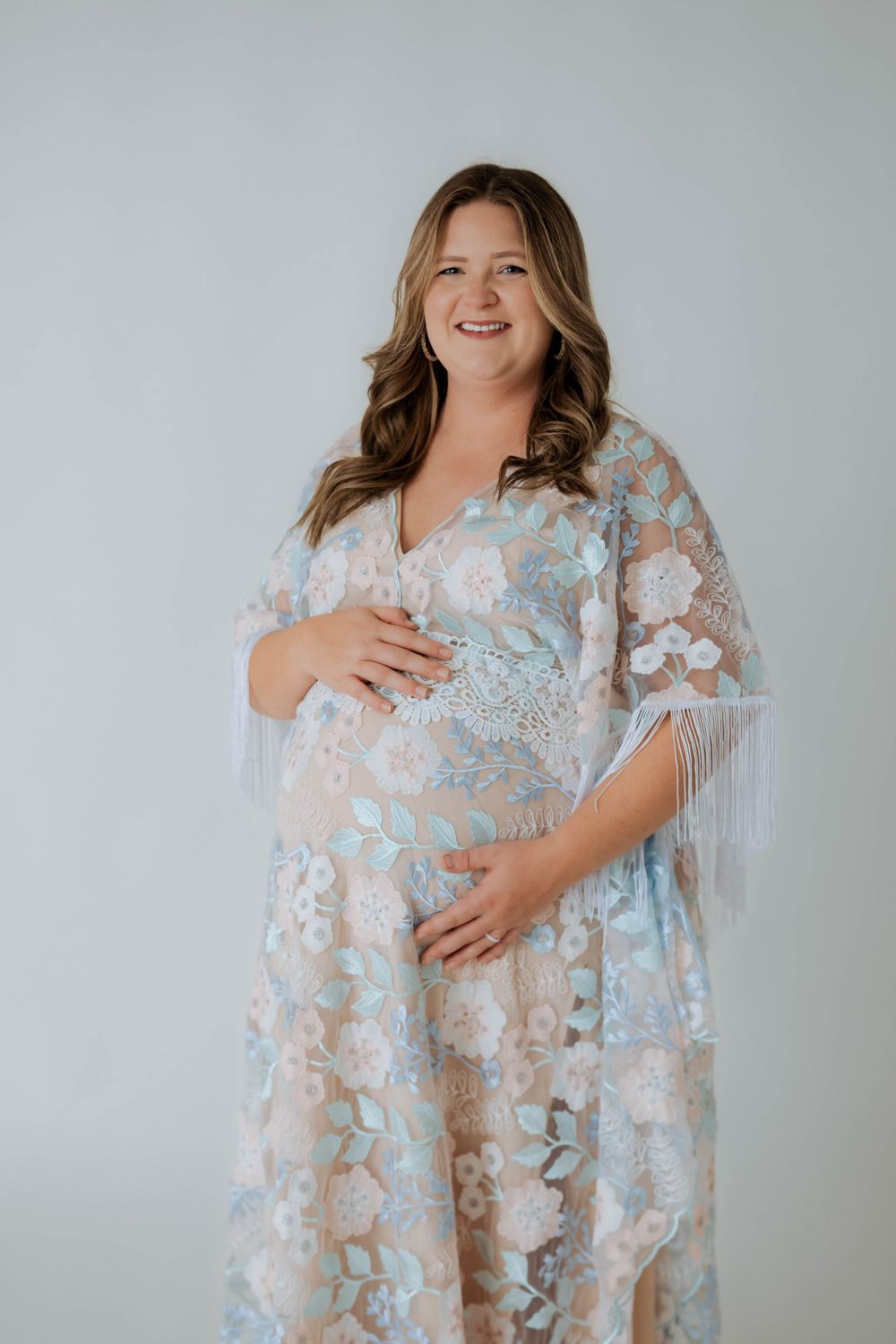 Expectant mother in a light blue and white lace maternity gown with flared sleeves poses against neutral background.