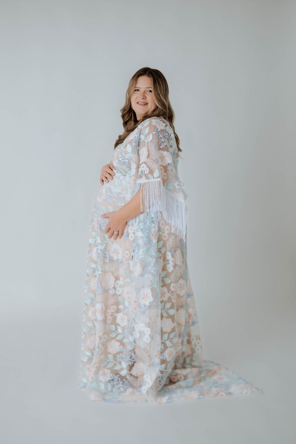 Side view of pregnant woman wearing flowing pastel floral dress with fringe detail against light grey backdrop.