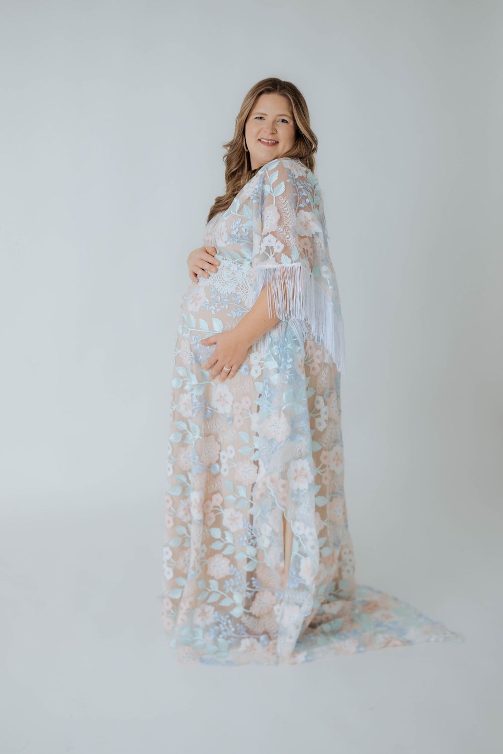Expectant mother wearing ethereal sheer floral robe in soft pastel colors poses against light grey backdrop for maternity photo.