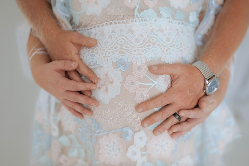 Close-up of hands with engagement ring resting on white lace dress showing intricate floral pattern details.
