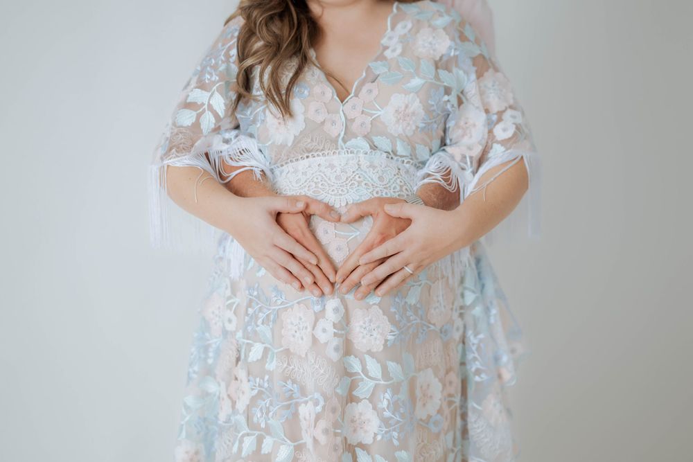 A delicate lace maternity dress in soft blue and white floral pattern is shown on a person making a heart shape with hands.