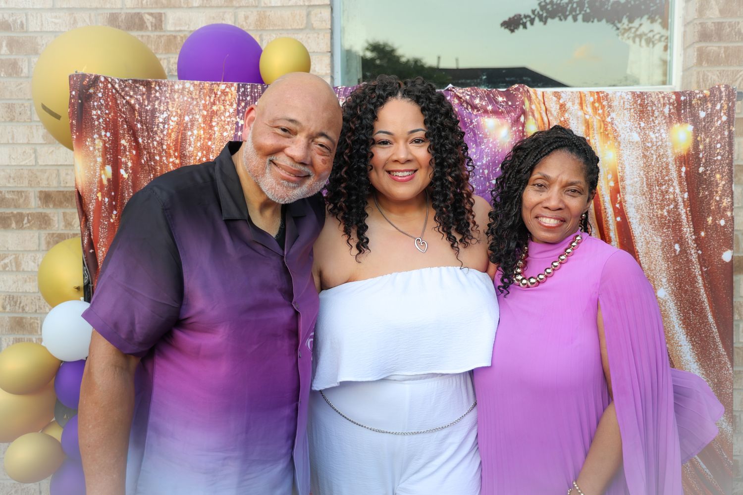 Three people smiling together at a festive celebration with purple and gold balloon decorations and a sparkly backdrop.