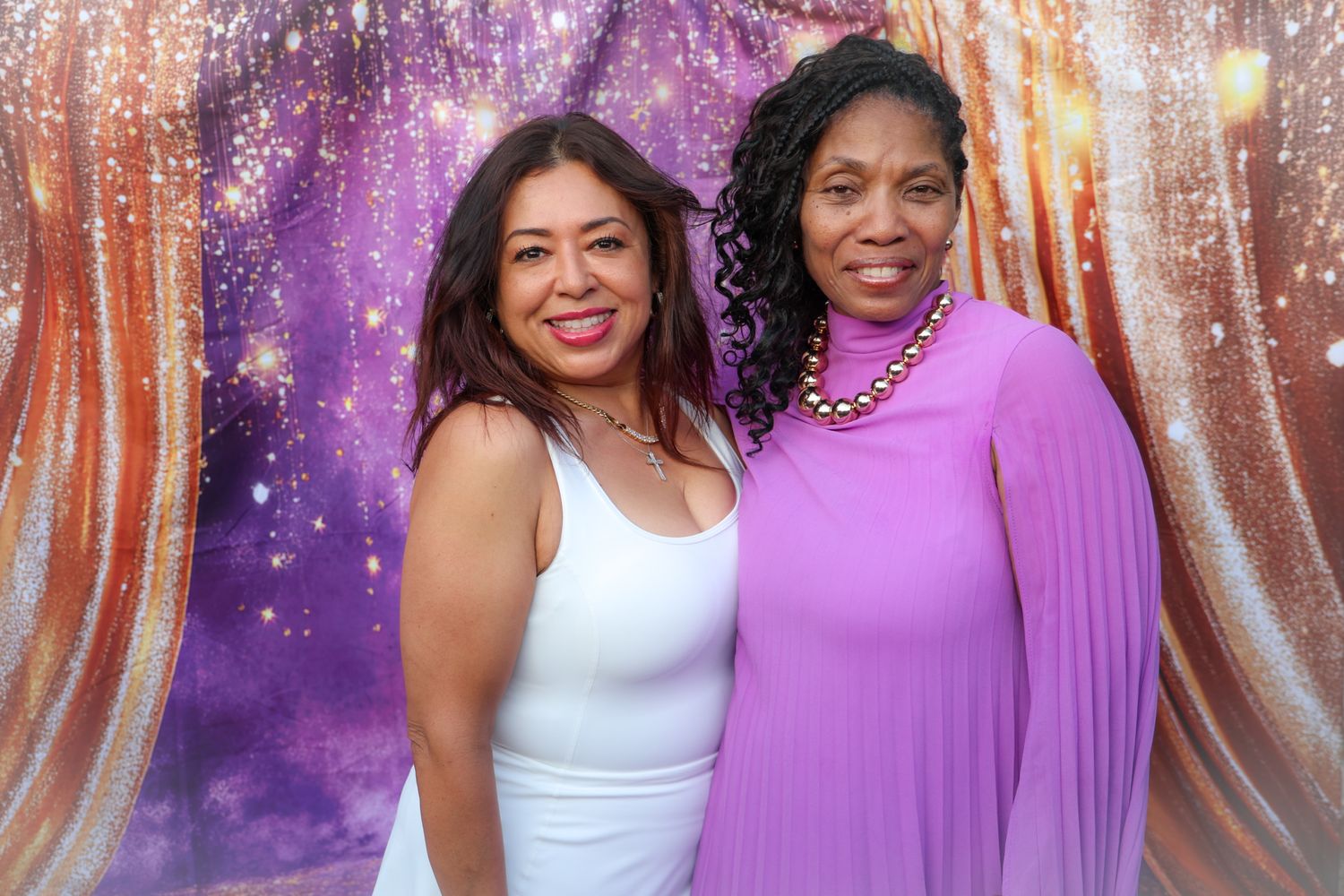 Two women smiling together in front of a purple and gold glittery backdrop at a formal event.