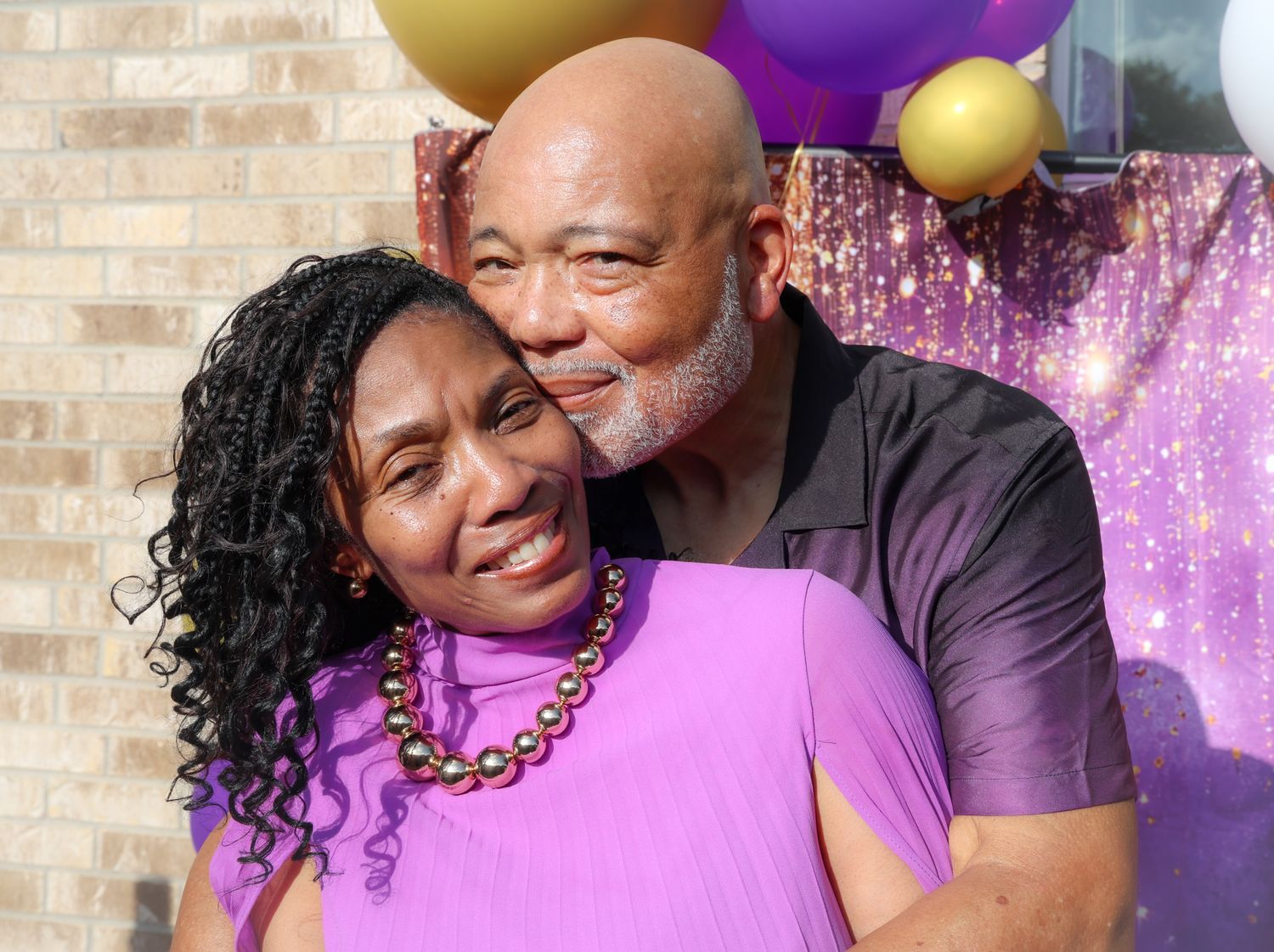 A joyful couple embraces outdoors, dressed in purple, with colorful balloons in the background.