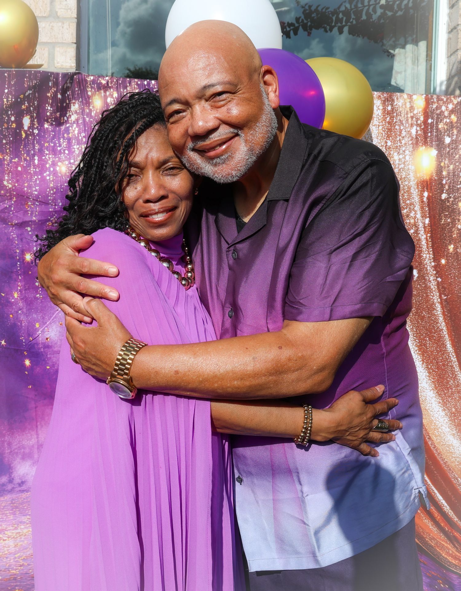 An elderly couple sharing a warm embrace, dressed in purple outfits, smiling joyfully in front of festive balloons.