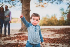 Toddler in striped shirt reaching up while playing in fallen autumn leaves.
