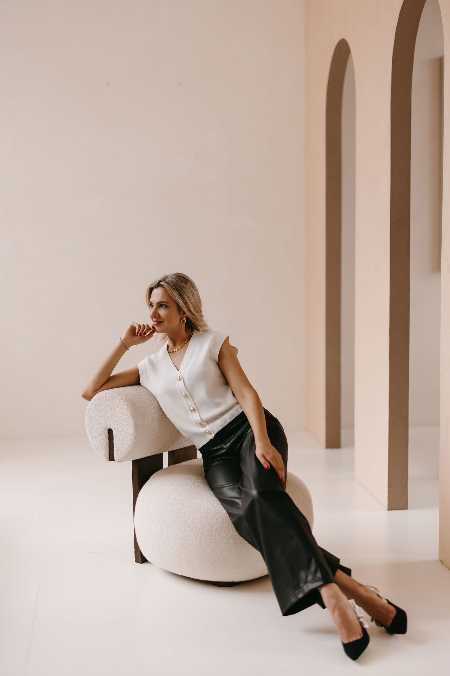 Model lounges on white curved chair wearing business casual outfit with black pants in minimalist architectural setting.