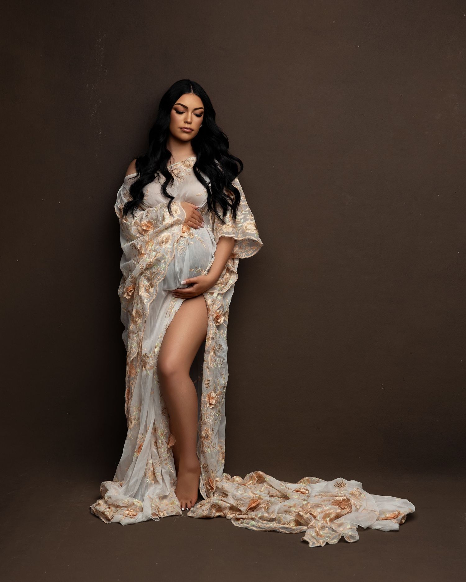 Elegant maternity portrait in flowing cream and gold robe against dark brown backdrop with dramatic studio lighting.