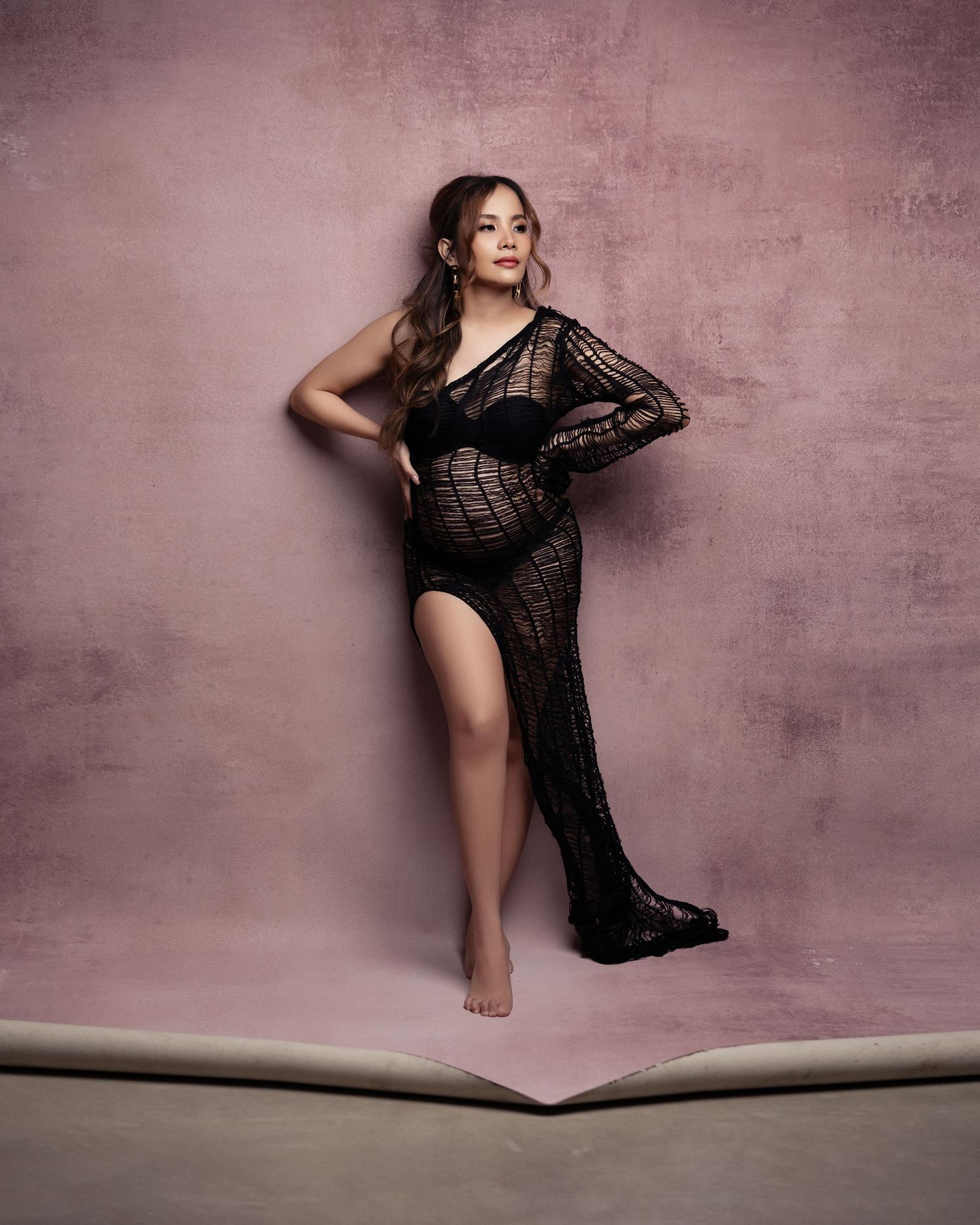 Elegant black lace maternity gown worn by a woman posing against a pink textured backdrop in a glamour photoshoot.