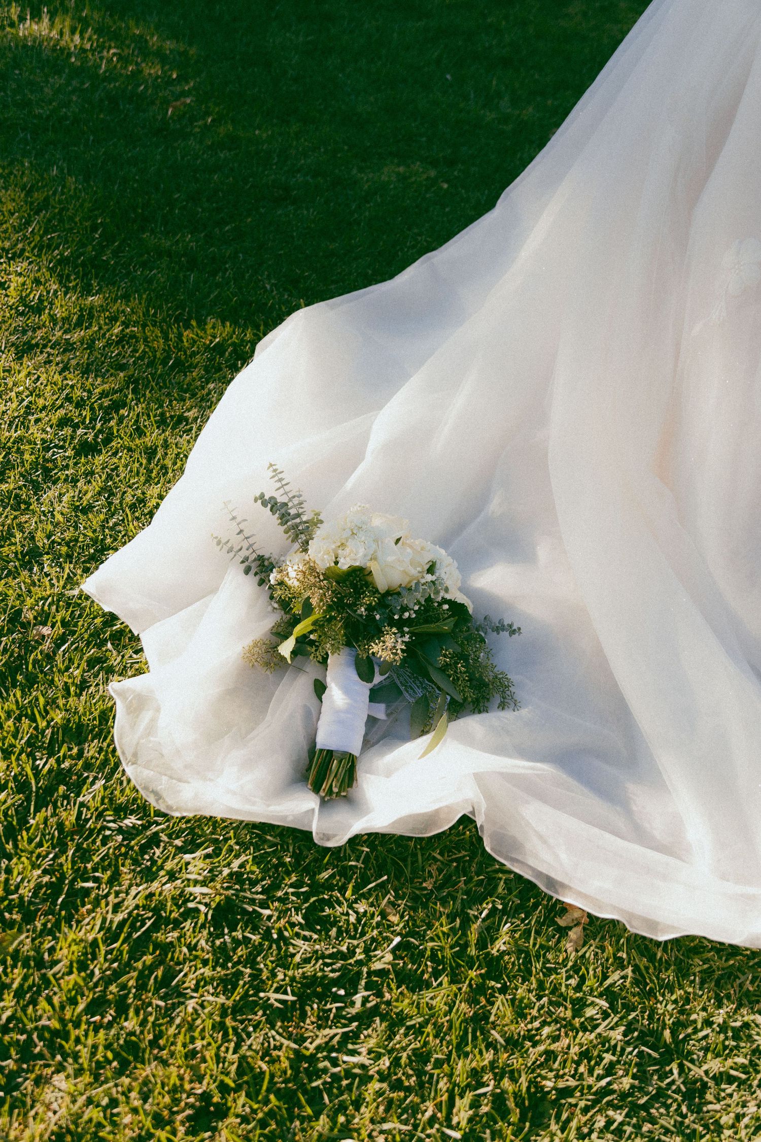 White wedding dress train with delicate floral bouquet trails gracefully across lush green grass in golden sunlight.