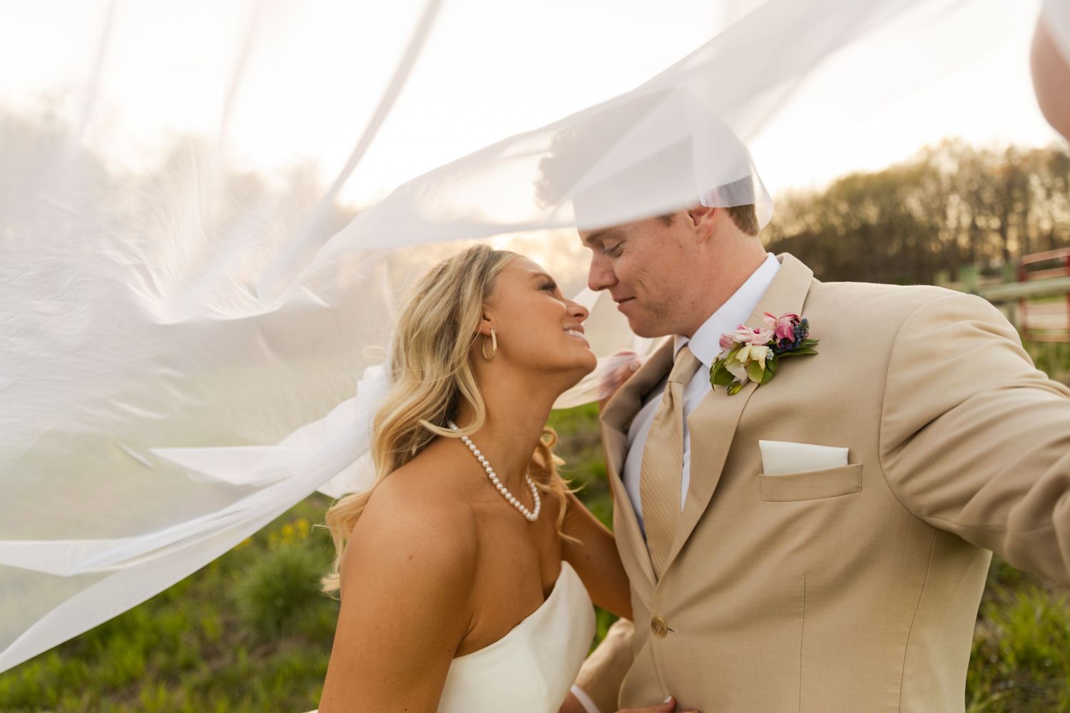 A romantic wedding moment with flowing veil and beige suit against a natural backdrop.