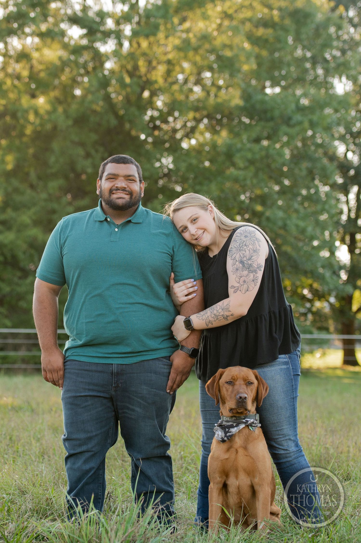 A couple poses with their brown dog in a grassy field during golden hour.