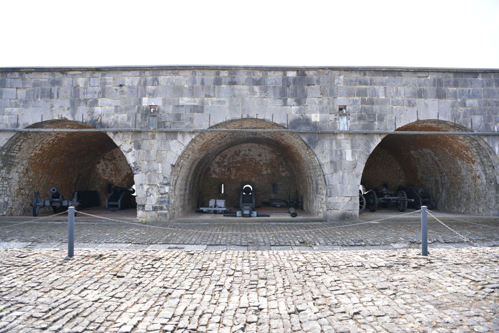 Three stone archways with cobblestone ground in front of an old fortress wall provide shelter for visitors.