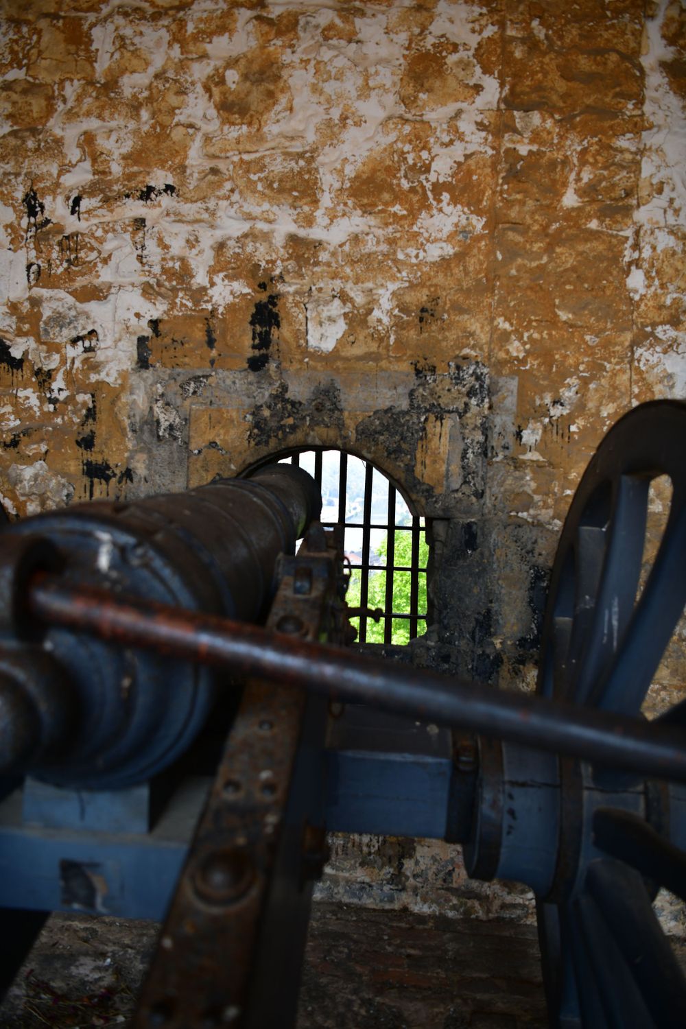 View through barred window of old fort or prison with weathered stone walls and rusty iron machinery in foreground.