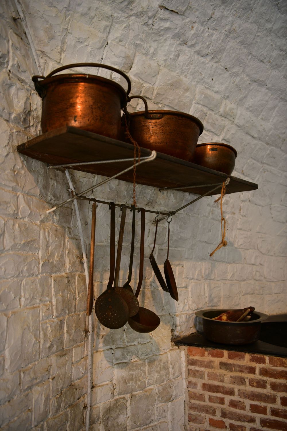 Vintage copper cooking pots and utensils hang on a rustic shelf against a white brick wall in an old kitchen.