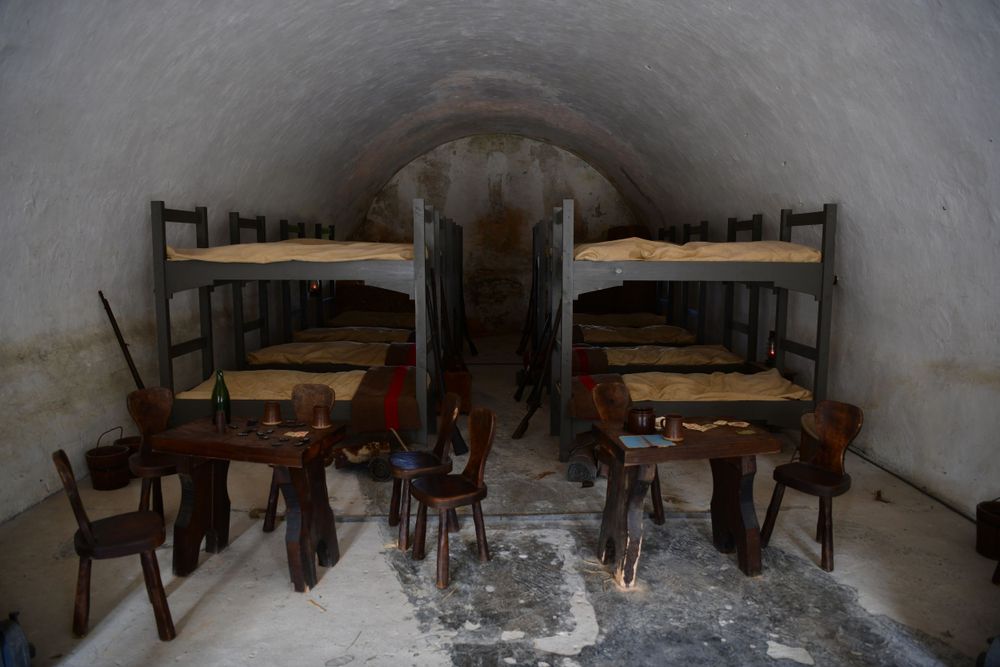 Historic prison cell interior with wooden bunks and chairs arranged along stone walls in a dimly lit vault-like space.