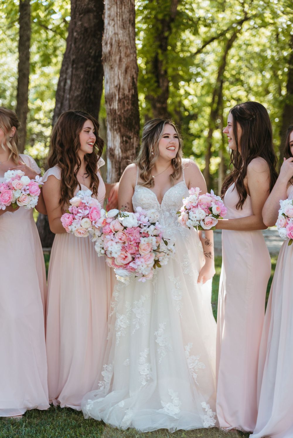 Wedding party in blush pink dresses and white gown pose with floral bouquets in front of trees.