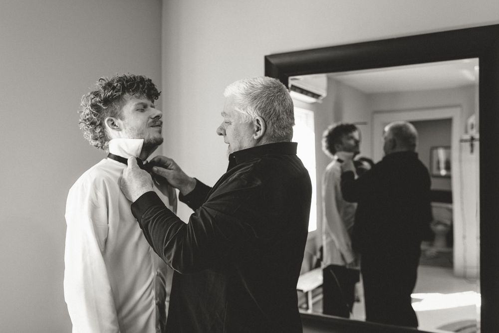Black and white image showing preparation moment with groom and father formal attire adjustments.