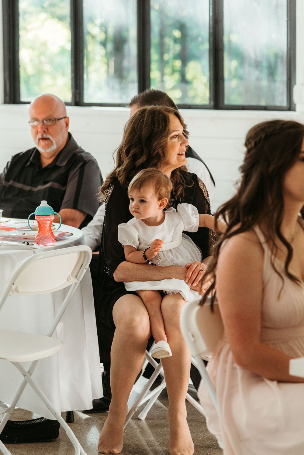 Wedding reception scene with guests seated at white tables in bright indoor venue.