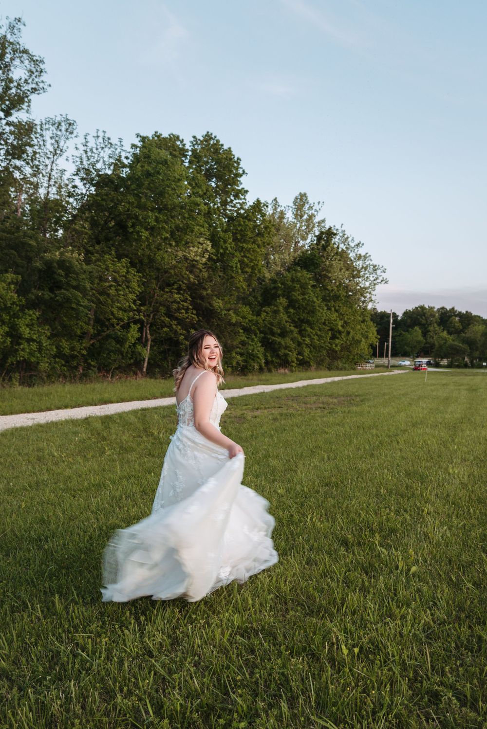 Bride in lace wedding dress twirls in flowing gown on grassy field with trees in background.