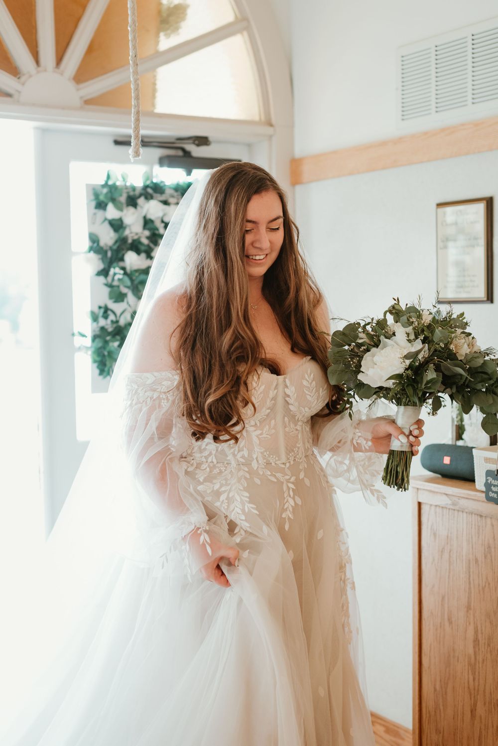 Bride holding bouquet poses in elegant lace wedding gown near arched window.