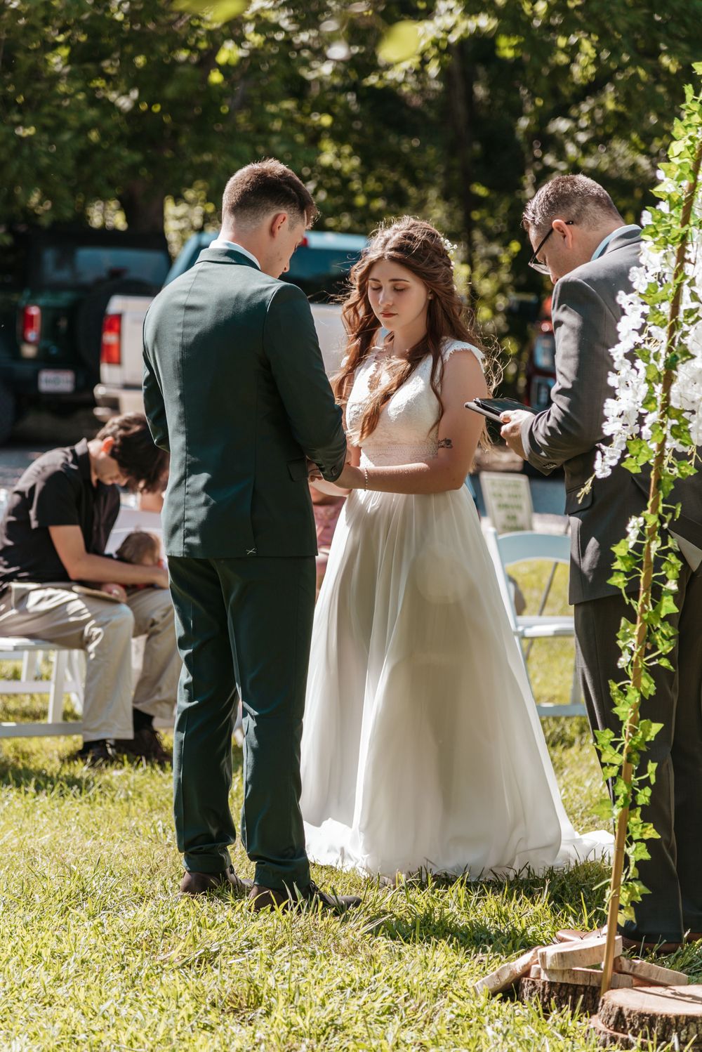 Wedding party gathered outdoors on a sunny day with decorative floral arch in background.