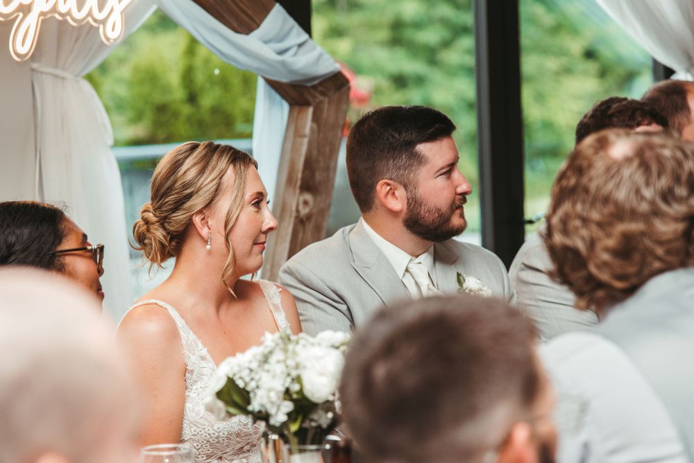 Wedding reception scene with guests mingling and conversing around decorated tables by large windows.